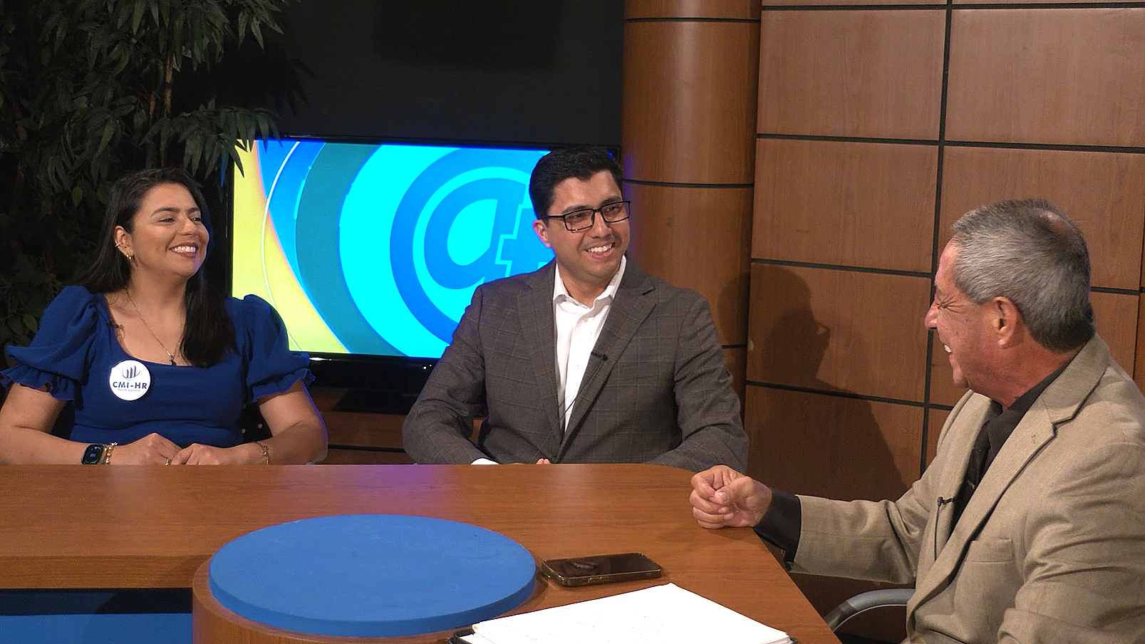 Three people seated at a news desk, with a blue and white graphic displayed on the screen behind them.