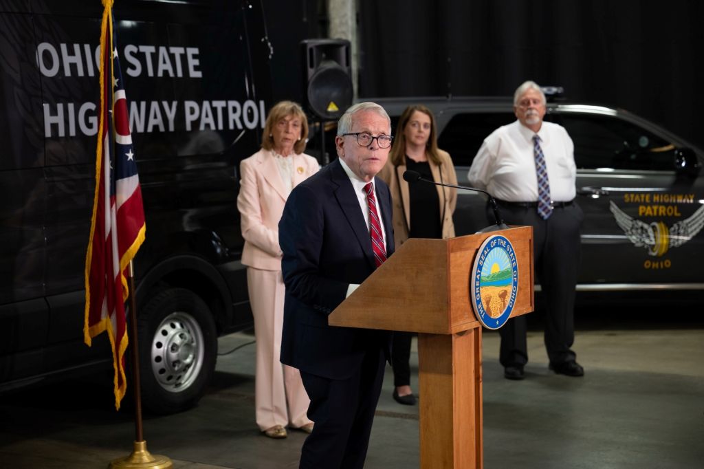 Ohio State Highway Patrol logo and vehicle visible behind a podium where a man in a suit is speaking at a press conference.