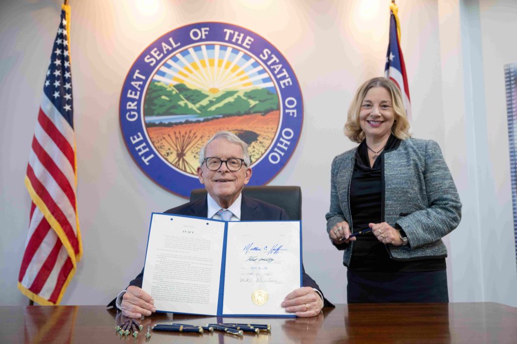 Two people standing in front of a seal of the state of Ohio, one holding a document and the other smiling.