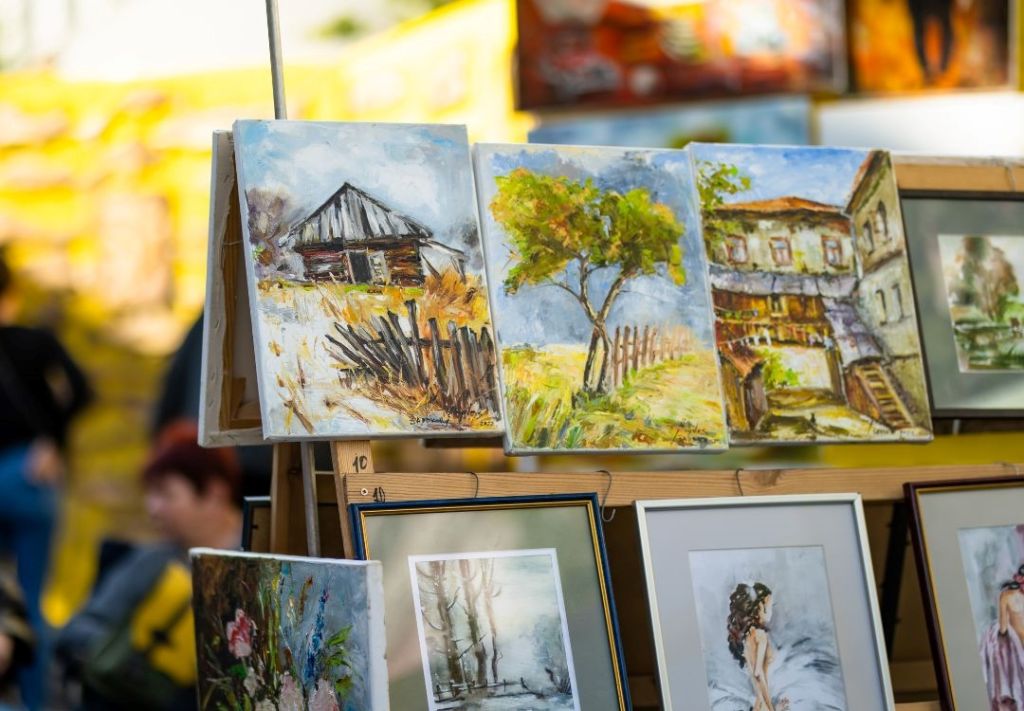 An outdoor art market display featuring several framed paintings depicting rural landscapes, buildings, and nature scenes.