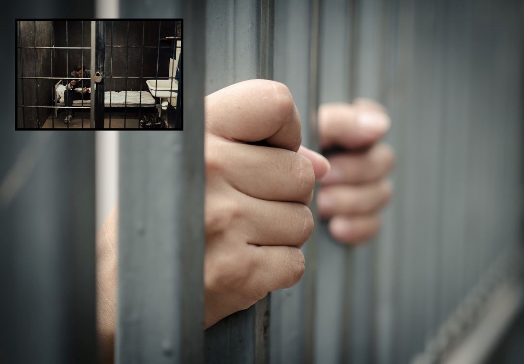Hands gripping metal bars of a prison cell, with a person visible through the window in the background.