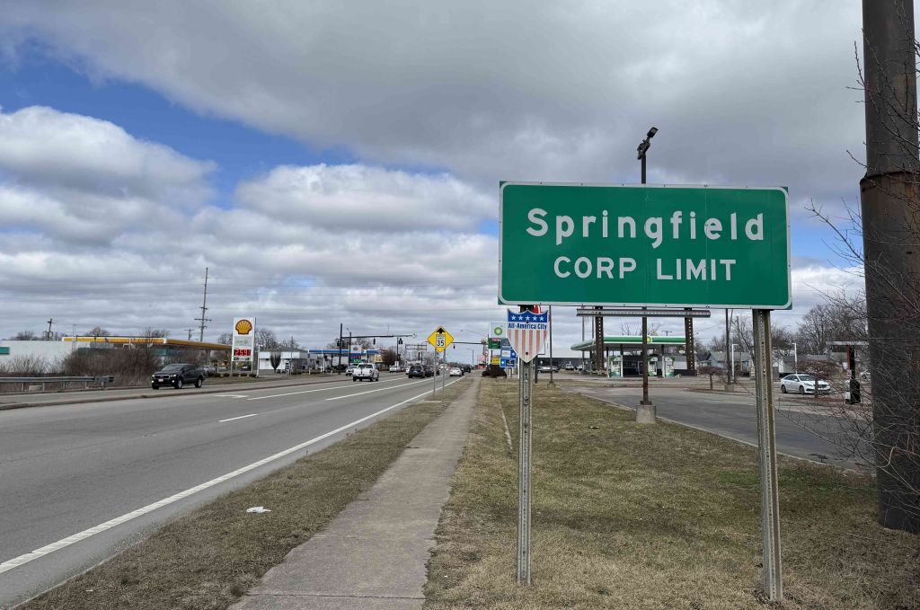 Springfield CORP LIMIT road sign on a highway with cars and buildings in the background.