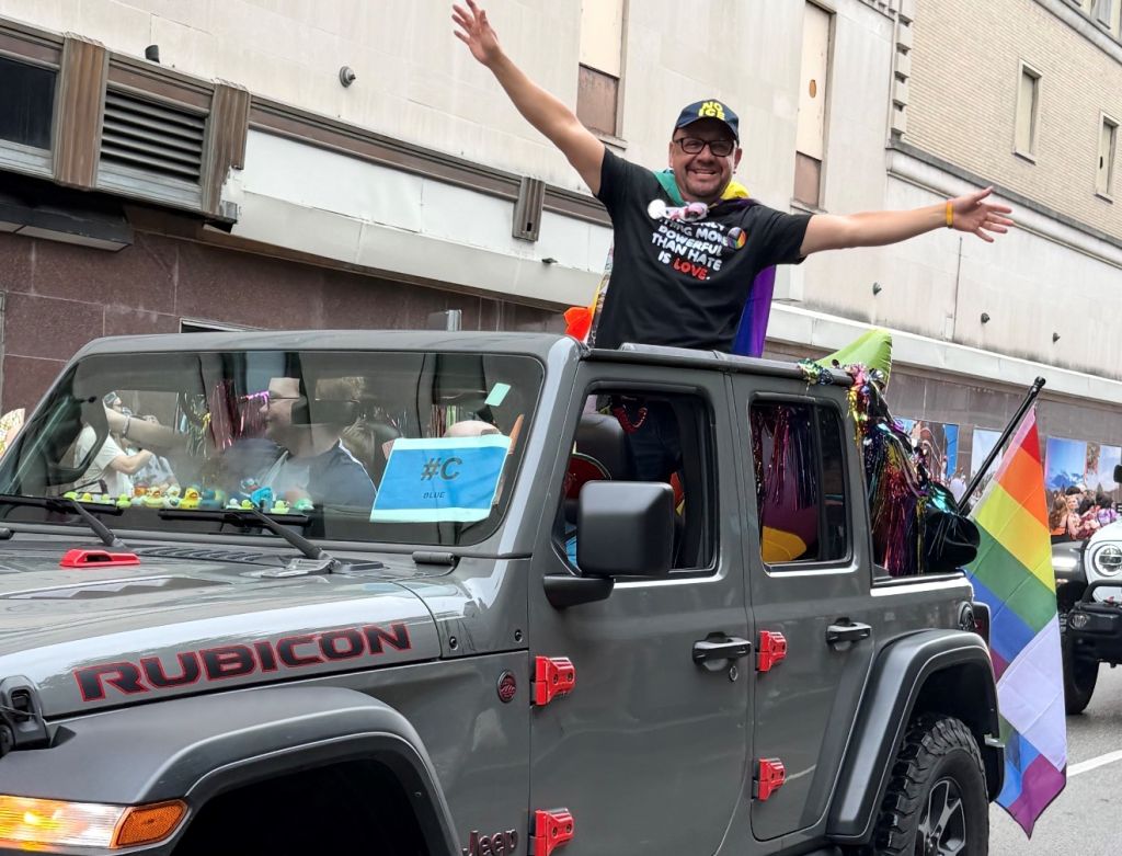 A person standing on the roof of a Jeep Rubicon waving their arms at a Pride parade, with colorful flags and decorations visible in the vehicle.