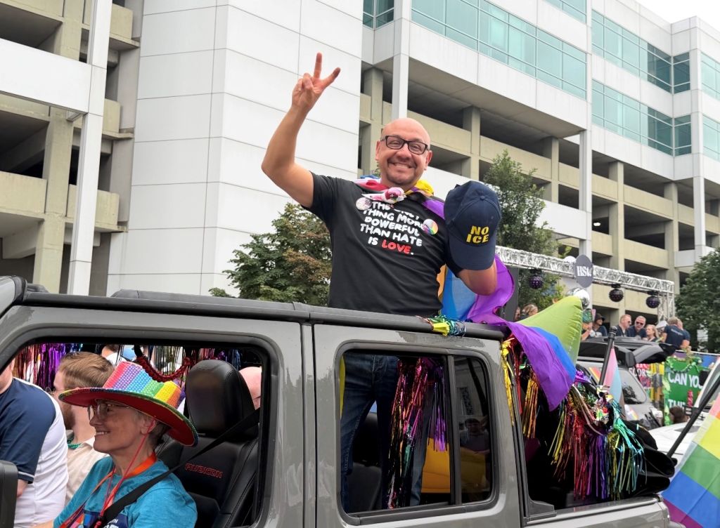 A smiling man wearing a "The love that dare not speak its name is love" t-shirt stands on top of a colorfully decorated vehicle during a parade or celebration.