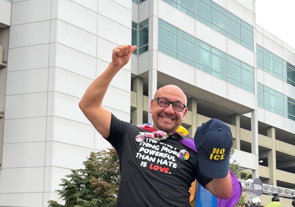 A smiling man wearing a shirt with the text "The thing most powerful than hate is love" and holding a "No ICE" sign in front of a modern glass building.