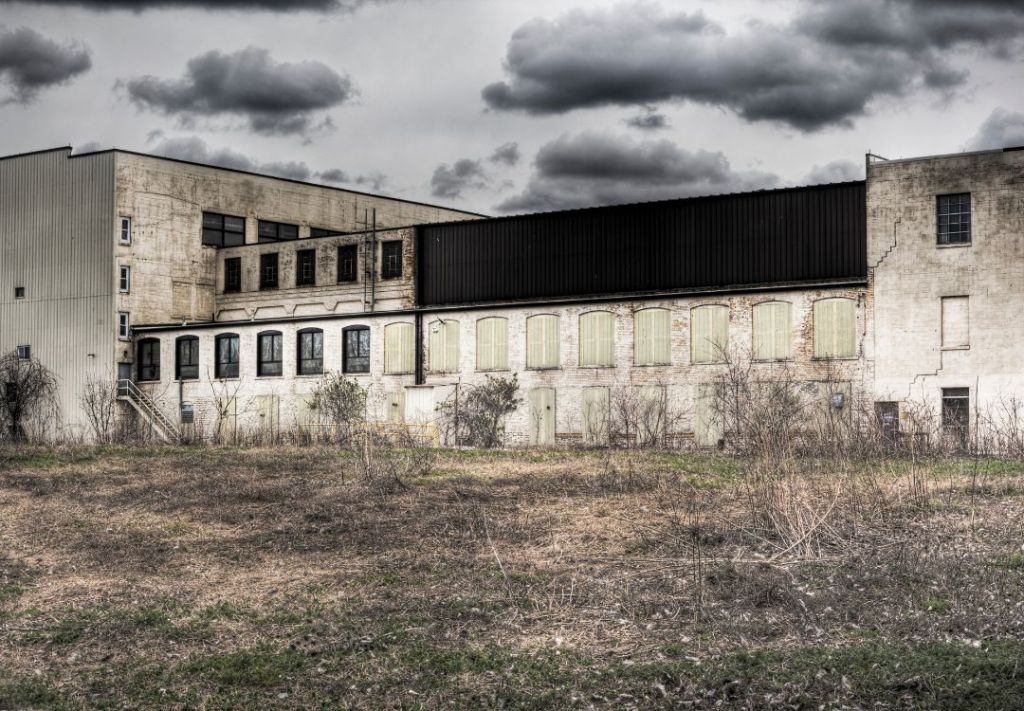 A dilapidated industrial building with boarded-up windows and a black metal roof stands in a grassy field under a cloudy sky.