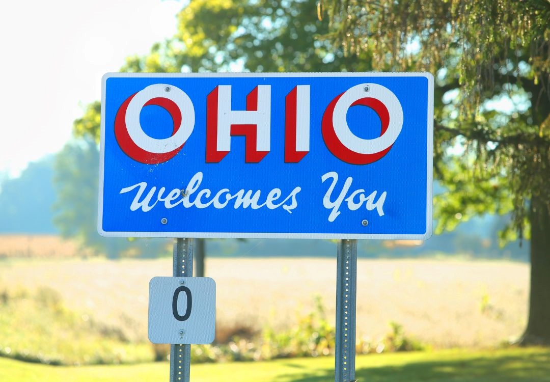 Blue and red "Ohio Welcomes You" road sign with the number "0" displayed below it, surrounded by trees and greenery.