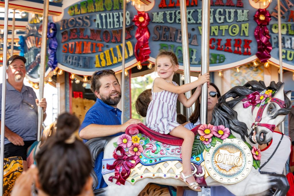 A colorful carousel with a variety of decorative horses and other carnival-themed elements. The carousel is surrounded by signs and banners with text including "Ross Athens Belnon Summit Ashland" and "Muskingum Clark Gum".