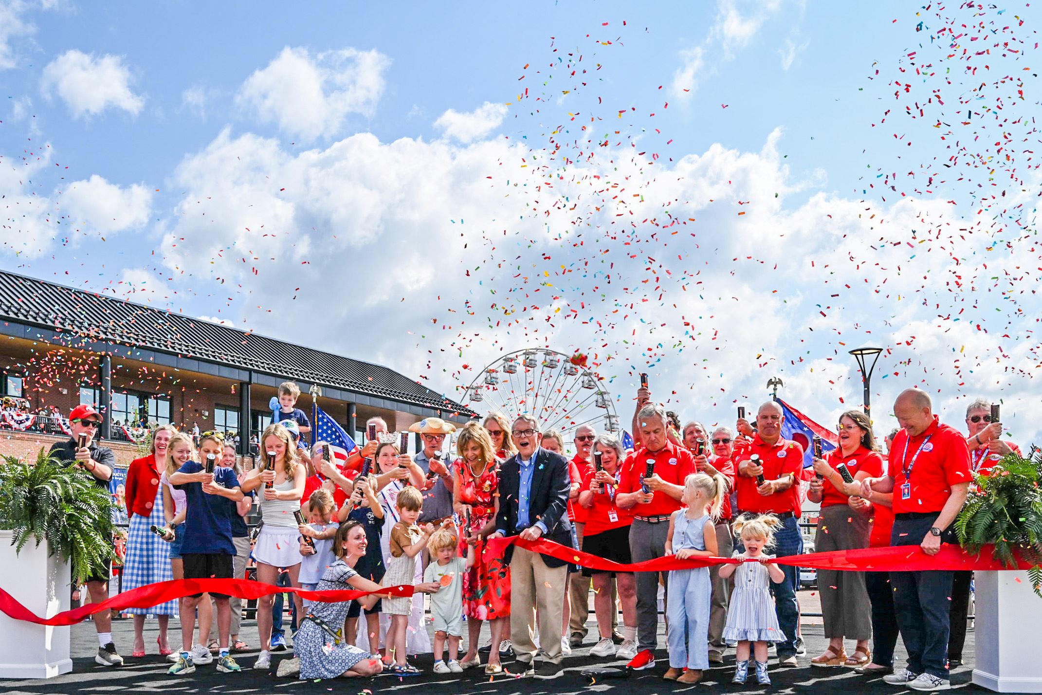 A crowd of people in red shirts and waving American flags gathers around a ribbon-cutting ceremony in front of a carnival-style building with a Ferris wheel.