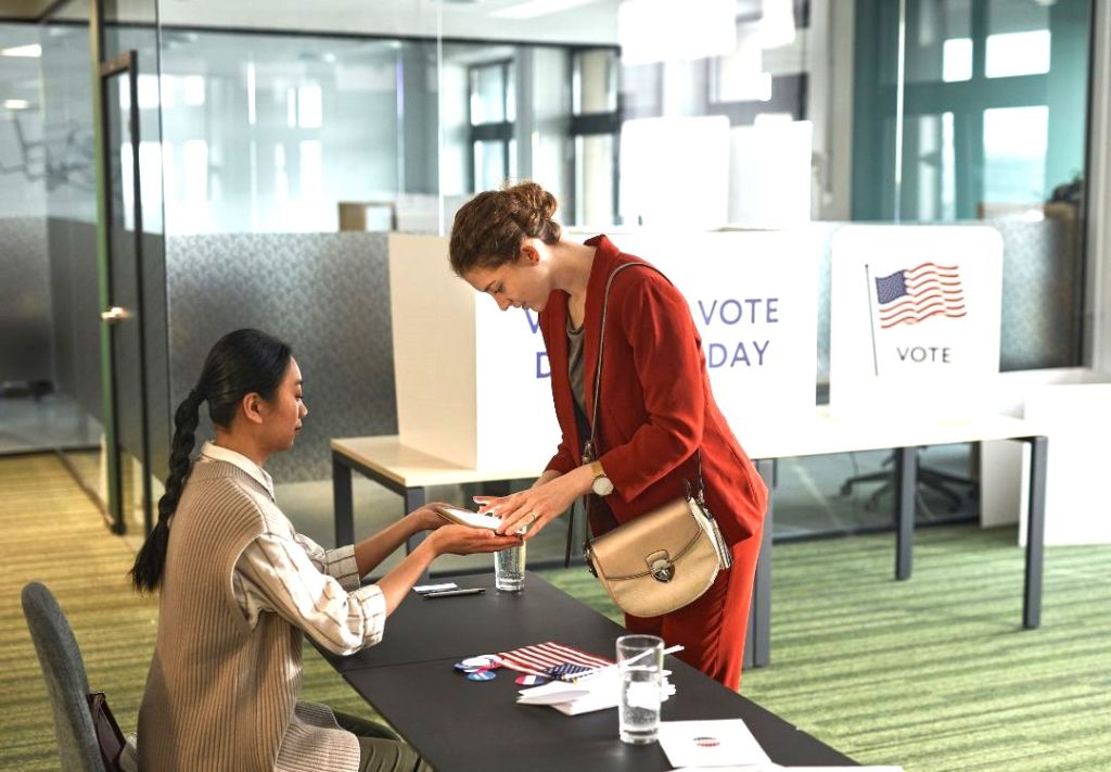 Two women at a voting booth, one in a red jacket handing something to the other woman seated at the table. A "Vote Today" sign and American flag are visible in the background.