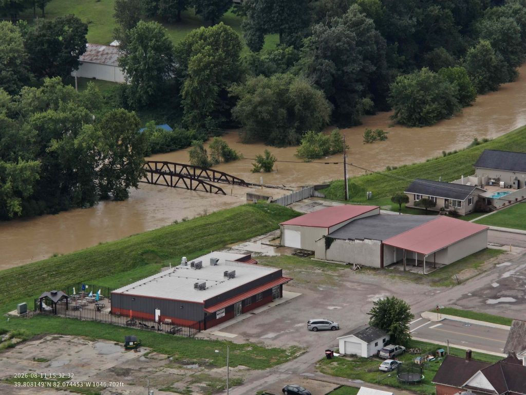 Aerial view of a flooded rural area with buildings, vehicles, and a damaged bridge over a swollen river.