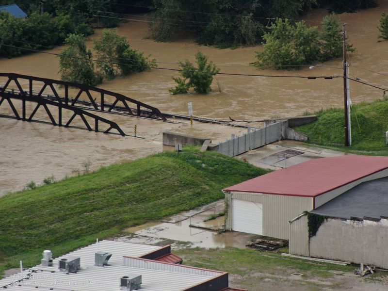 A flooded river flows under a damaged bridge, with buildings and power lines visible on the banks.