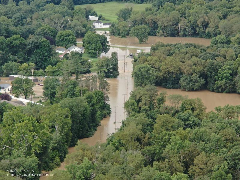 Aerial view of a flooded rural area with a submerged road and surrounding trees and buildings.