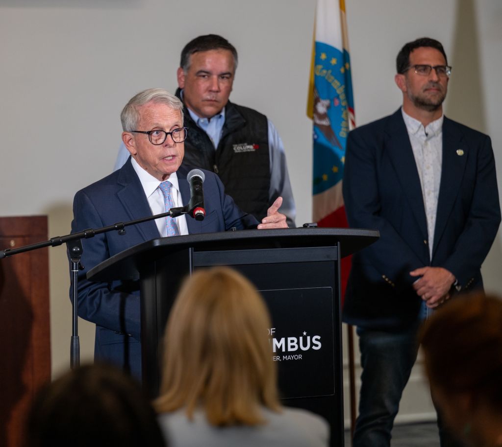 Three men, including the mayor, speaking at a podium with "MAYOR OF COLUMBUS" displayed.
