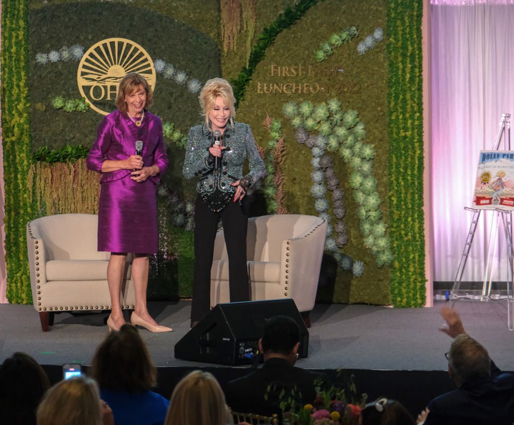 Two women on stage, one in a purple dress and one in a black and white outfit, speaking into microphones in front of a floral backdrop with the text "First Ever Luncheon" visible.